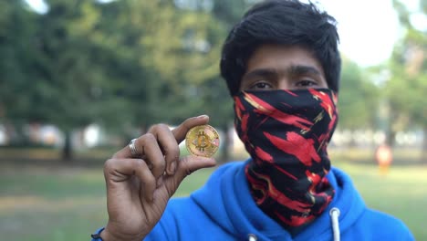 Ein-Indischer-Junge-In-Blauem-T-shirt-Und-Marke,-Die-Eine-Münze-Gold-Bitcoin-Ethereum-Outdoor-Park-Hintergrund-Hält