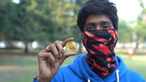 Close-up-shot-of-an-Indian-boy-in-blue-t-shirt-and-mark-holding-a-coin-gold-bitcoin-Ethereum-outdoor-park-background