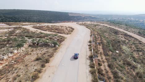Aerial-drone-shot-following-a-black-car-through-a-spruce-forest-trees-on-hilly-terrain-at-daytime-in-Katzir,-Israel