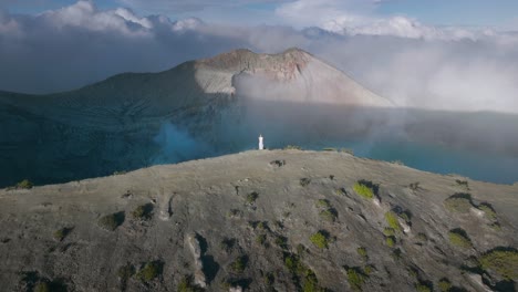 Woman-in-white-dress-standing-on-cliff-edge-overlooking-volcano-with-blue-water-lake,-aerial