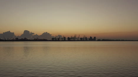 Aerial-view-moving-towards-shipping-port-cranes-with-silhouette-tide-marker-on-Miami-coastline-at-sunset