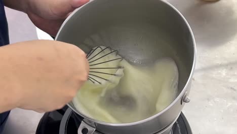 Close-up-shot-of-a-professional-pastry-chef-hand-mixing-and-whisking-cooked-egg-white-with-steaming-hot-water-on-electric-cooktop-in-a-commercial-bakery-setting