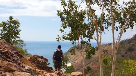 Man-hiking-through-wilderness-outdoors-in-the-hills-with-rocky-terrain-overlooking-beautiful-blue-ocean-on-a-remote-tropical-island-destination,-walking-away-from-camera-towards-sea-view