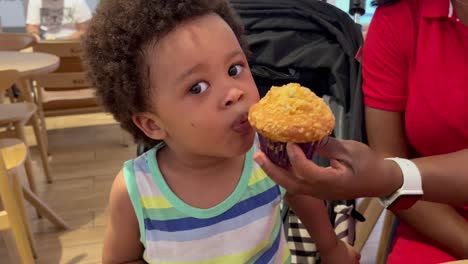 Entzückendes-Und-Exotisches-Zweijähriges-Afroeuropäisches-Kind,-Das-Neben-Seiner-Mutter-In-Einer-Cafeteria-Einen-Köstlichen-Muffin-Isst