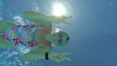 Cheeky-Female-Athlete-Swimmer-Dives-Down-To-Underwater-Camera,-Smiles-And-Gives-Peace-Sign,-4K-Slow-Motion