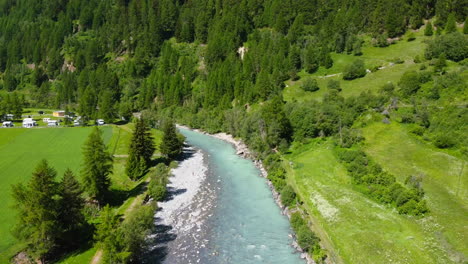 Beautiful-blue-river-surrounded-by-vibrant-green-trees-on-summer-day-in-Zernez-Switzerland,-aerial