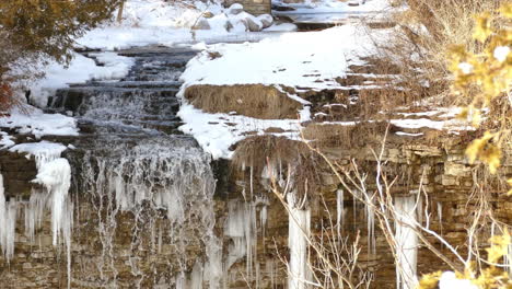 Small-waterfall-stream-falling-down-rocky-cliff-in-winter-forest,-pan-left-view