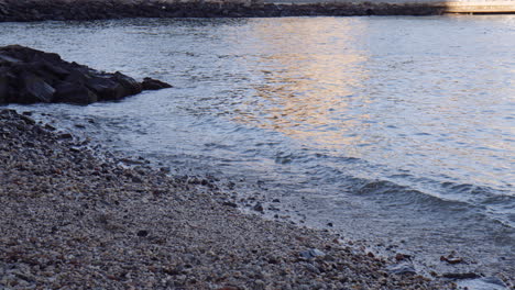 Ocean-waves-buffet-shady-pebble-beach-with-reflection-of-Brooklyn-Bridge,-handheld