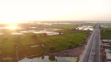 Vista-Aérea-De-La-Puesta-De-Sol-Amarilla-Dorada-En-El-Horizonte-Sobre-Campos-Verdes-Inundados-En-Sindh