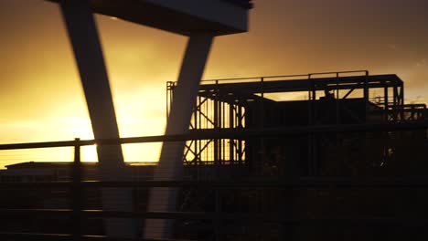 Looking-Out-Car-Window-Going-Past-Sillhouute-Construction-Site-During-Golden-Yellow-Sunset
