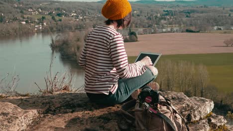 Close-up-with-tracking-shot-of-a-woman-who-opens-the-screen-of-her-laptop-on-the-edge-of-a-cliff,-she-looks-at-the-landscape,-a-camera-is-placed-on-her-backpack