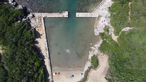People-swimming-near-ancient-Roman-port-in-Italy,-aerial-top-down-ascend-view