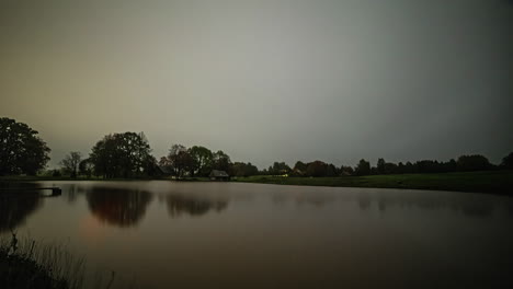 Timelapse-Of-Lake-House-With-Lights-Turning-On-And-Off-From-Across-Calm-Reflective-Lake-At-Night-With-Grey-Clouds,-Static-Shot