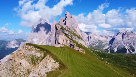 Pull-back-aerial-view-from-of-the-peaks-of-Seceda-with-green-pastures,-and-hiking-trails-in-the-foreground,-and-mountain-peaks-in-the-background-in-the-Italian-Dolomites-in-South-Tyrol,-Italy