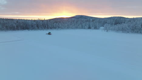 Tractor-travelling-towards-snowy-Norbotten-woodland-preparing-Lapland-ice-race-track-at-sunrise-aerial