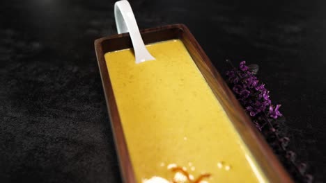 Creamy-Pumpkin-Soup-in-a-Wooden-Square-Bowl-Decorated-with-Flower-and-Herbs-on-a-Black-Table---Revealing-dolly-shot
