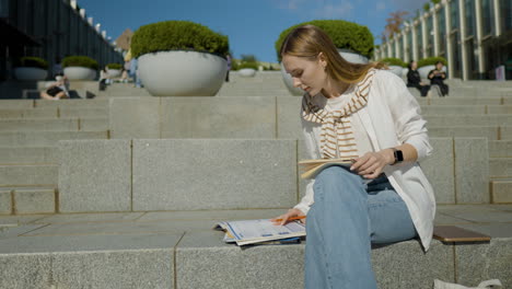 Schöne-Studentin,-Die-Auf-Der-Treppe-Im-Freien-Sitzt-Und-Notizen-Auf-Einem-Notizbuch-Auf-Dem-Universitätscampus-Schreibt