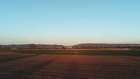 Zooming-out-of-landscape-over-agricultural-field-during-sunset-with-full-moon-on-sky-in-Germany,-Europe