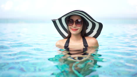 Pretty-Asian-woman-wearing-a-large-floppy-black-and-white-striped-sun-hat-and-sunglasses-to-protect-her-face-enjoys-the-warm-water-of-her-resort's-infinity-pool