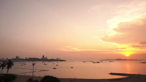 Ein-Atemberaubender-Sonnenuntergang-In-Goldrosa-über-Der-Bucht-Von-Pattaya-Erleuchtet-Angedockte-Boote,-Die-Auf-Ruhigem-Wasser-Schwimmen,-Während-Straßenlaternen-Zu-Leuchten-Beginnen