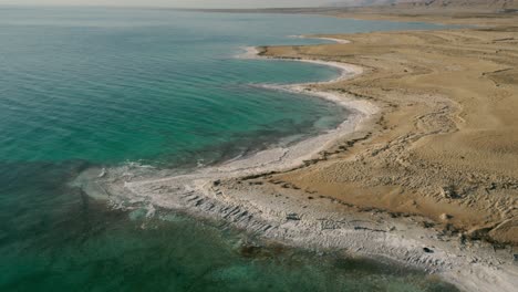 Aerial,-arid-and-barren-coast-with-calm-clear-teal-blue-water,-wide-shot