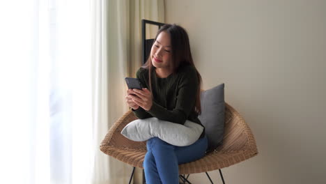 pretty-brunette-sitting-in-a-wicker-chair-wearing-a-dark-green-shirt-and-blue-jeans-with-a-white-pillow-in-her-lap-smiles-as-she-text's-on-her-cellphone-with-brilliant-light-coming-through-the-window