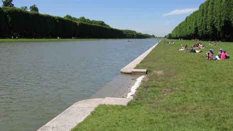 People-resting-on-a-lakeside,-in-the-gardens-of-the-Versailles-Palace,-Paris,-France