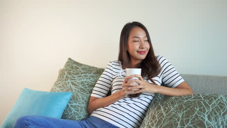 attractive-brunette-woman-drinking-from-a-white-mug-wearing-a-striped-white-shirt-and-blue-jeans-leans-back-on-a-couch-with-green-and-blue-pillows-and-smiling-contently