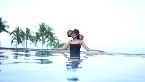 A-young-Asian-woman-wearing-a-black-hat-and-camisole-relaxes-at-the-edge-of-an-infinity-pool-while-enjoying-the-sunny-tropical-outdoors