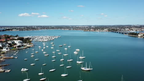 Aerial:-Drone-flying-over-a-bay-of-boats-tracking-along-the-coastline-to-reveal-a-marina-and-bridge-in-the-distance,-in-Sydney-NSW