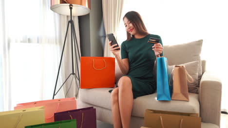 Stilvolle-Asiatische-Shopaholic-Frau,-Die-Kreditkartennummern-Auf-Dem-Smartphone-Eingibt,-Während-Sie-Auf-Dem-Sofa-Im-Wohnzimmer-Sitzt