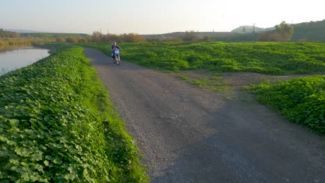 Person-rides-motorbike-on-gravel-road-next-to-stream-and-lush-vegetation,-medium-shot