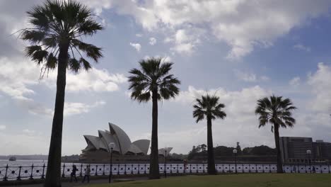 Sydney-Opera-House,-Sydney,-Nueva-Gales-Del-Sur,-Australia---Gente-Caminando-En-El-Parque-Con-Palmeras-Cerca-De-La-ópera-Bajo-El-Cielo-Azul-Y-Nubes-Blancas---Gran-Plano