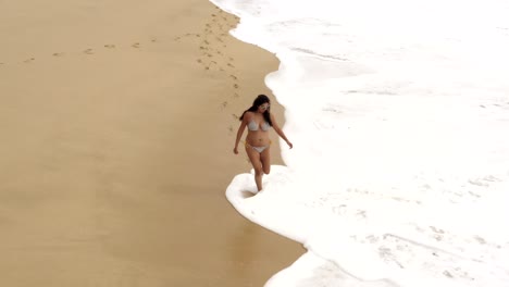 Beautiful-tanned-female-in-swimwear-walking-along-beach-in-waves,-aerial-view