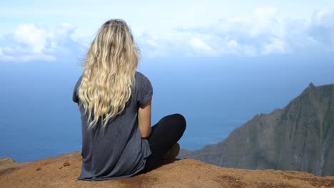 A-blonde-young-woman-sitting-on-cliff-in-the-Napali-Coast-State-Park-in-Kauai,-Hawaii
