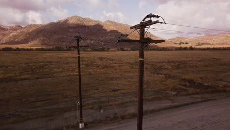 Power-lines-over-the-dusty-road-in-the-desert,-parched-land-and-the-mountains-in-the-background