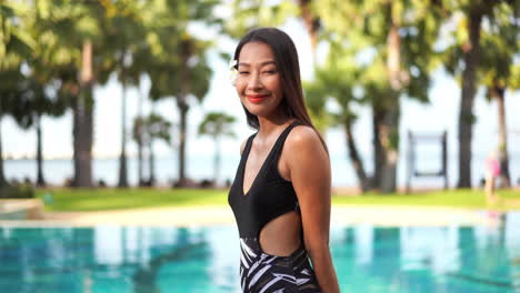 Beautiful,-shy-Asian-girl-with-black-bathing-suit-smiling-to-the-camera,-blurred-swimming-pool-background