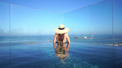 A-woman-stands-waist-deep,-in-the-waters-of-a-resort-infinity-pool-looking-out-through-the-glass-barrier-to-the-ocean-waters-beyond