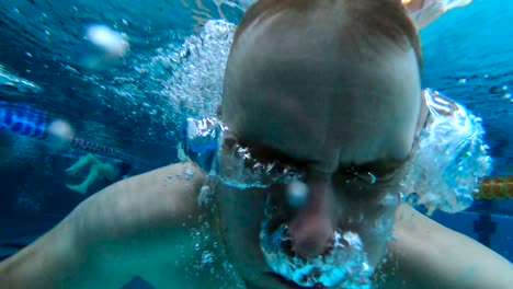 Ein-Mann-Schwimmt-In-Einem-Hallenbad,-Während-Er-Direkt-In-Die-Kamera-Schaut,-Dann-Taucht-Und-Luft-Aus-Der-Nase-Bläst