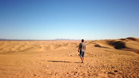 Hombre-Solitario-Parado-En-La-Arena-Del-Desierto-Y-Mirando-Las-Dunas-Y-El-Horizonte-Del-Cielo-Despejado