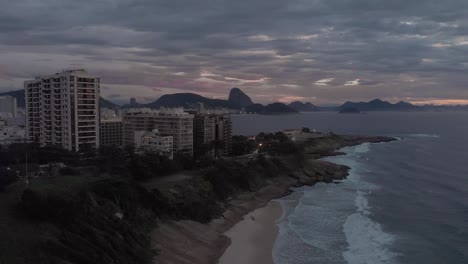 Aerial-descend-showing-the-Copacabana-fort-behind-a-small-beach-with-cliffs-in-the-foreground-and-the-Sugarloaf-mountain-in-the-background-on-an-overcast-day-at-sunrise-in-Rio-de-Janeiro