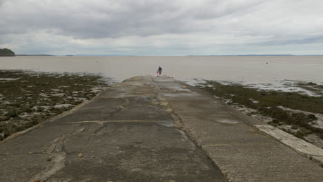 Grandparent-and-grandchild-explore-coastal-slipway-edge-watching-the-sea-on-cloudy-day,-wide-shot
