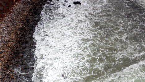 Aerial-fly-over-of-rocky-beach-with-grey-waves-crashing-onto-shore