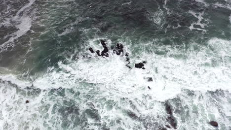 Dark-ocean-waves-crashing-into-white-foam-on-black-rocks,-Aerial-View
