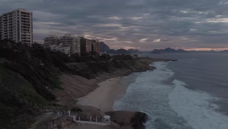 Early-morning-sunrise-aerial-descend-with-waves-coming-in-on-the-cliffs-and-revealing-the-pristine-sand-of-the-Devils-beach-in-Rio-de-Janeiro-with-an-orange-glow-at-the-horizon