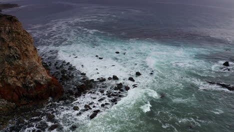 White-ocean-waves-crashing-at-rocky-foot-of-steep-cliff,-Aerial