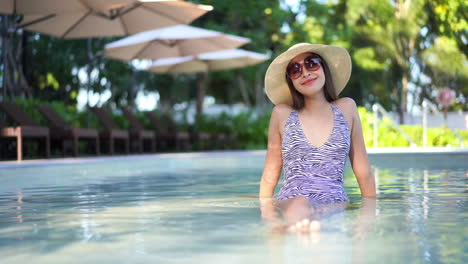 A-young-Asian-woman,-wearing-a-black-and-white-patterned-one-piece-bathing-suit,-floppy-sun-hat,-and-designer-sunglasses,-lounges-in-the-shallow-waters-of-a-resort-pool