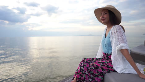 An-attractive-young-Asian-woman-in-a-colorful-printed-flowing-shirt-sits-on-a-cushioned-seaside-seat-looking-out-on-the-ocean