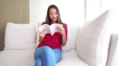 Asian-woman-is-comfortably-sitting-on-the-sofa-and-reading-a-book-journal-with-white-cover