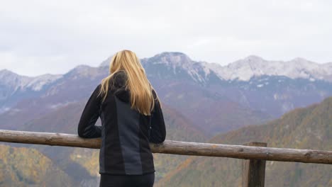 Blonde-young-woman-leaning-on-wooden-fence-looking-towards-mountains